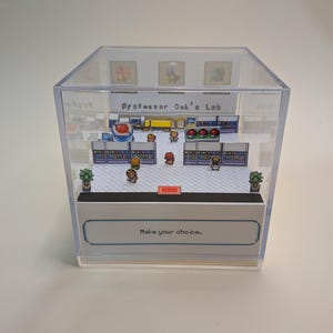 Può includere: Un diorama in cubo acrilico trasparente che presenta una scena in miniatura 2D del laboratorio del Professor Oak tratto da un videogioco. L'interno mostra personaggi pixelati, scaffali e tre Poké Ball. Il testo recita Professor Oak's Lab e Make your choice.