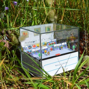 May include: A clear acrylic cube diorama featuring a miniature paper-crafted scene titled Professor Oak's Lab. The interior displays small character figures, laboratory equipment, and shelves. The base includes the printed text Role your choices.