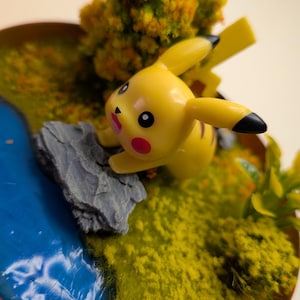 Op de afbeelding: Een geel Pikachu-figuurtje met zwartgepuntte oren en rode wangen, geplaatst op een grijze rots in een miniatuur diorama. De scène bevat een blauwe harsstroom, groene textuurgrond en een kleine boom met geel en groen gebladerte.