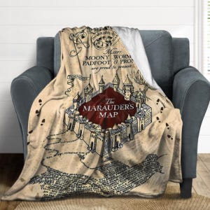 Puede incluir: Una manta beige con un estampado gráfico del Mapa del Merodeador de Harry Potter. El diseño incluye un escudo central de color rojo oscuro con el texto The Marauder's Map, rodeado de ilustraciones de castillos, huellas y caligrafía.