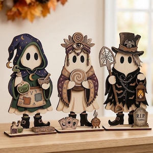 Puede incluir: Tres figuras de fantasmas de madera con ilustraciones detalladas. La primera lleva una capa morada con capucha, sosteniendo una botella de poción y un mortero. La segunda lleva un tocado y sostiene una bola de cristal. La tercera lleva un sombrero de copa, una pala y una linterna.