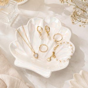 Könnte beinhalten: Eine weiße, irisierende Schmuckschale in Muschelform mit einer goldenen Halskette mit Herzanhänger, zwei goldenen Creolen mit Perlenanhängern, einem goldenen Ring, einem schlichten Ring und einem goldenen Armband mit kleinen weißen Perlen und Muschel.