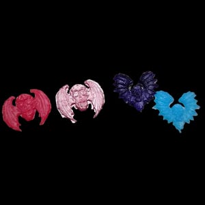 May include: Four small decorative pins shaped like winged skulls, arranged in a horizontal row. The pins are colored in shades of bright red, light pink, dark purple, and vibrant blue, each featuring detailed wing structures and skull faces.