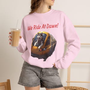 May include: A light pink long-sleeve sweatshirt featuring a graphic of a husky dog leaning out of the front of a yellow train. Above the train, the text reads "We Ride At Dawn!" in red letters. The sweatshirt has a crew neckline and long sleeves.