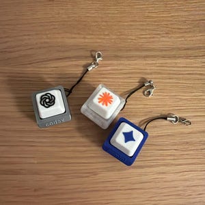May include: Three square mechanical keyboard keycap charms with black lanyards and metal clasps. One has a grey base with a black spiral, one a translucent base with an orange star, and one a blue base with a blue diamond shape. The word cod6x is on the grey base.