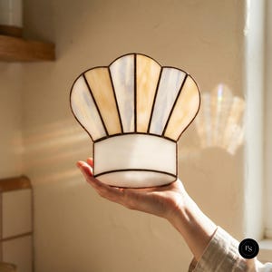Puede incluir: Un atrapasoles de vidrieras con forma de gorro de chef, que presenta paneles alternos de vidrio crema translúcido y blanco iridiscente unidos por líneas de soldadura de metal oscuro, diseñado para captar la luz y crear un brillo suave.