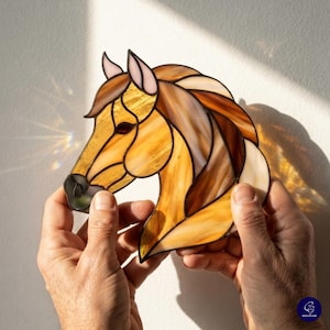May include: A stained glass suncatcher shaped like a horse head, featuring various shades of amber, tan, and brown glass pieces joined by dark metal solder lines. The design includes a light pink ear and a dark glass eye, creating a horse profile.