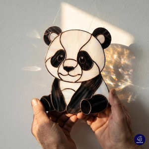 Puede incluir: Un atrapasoles de vidrieras con forma de oso panda sentado, compuesto por piezas de vidrio blanco opaco y negro translúcido unidas por líneas de soldadura de metal oscuro. El diseño incluye orejas redondeadas, manchas oculares y nariz.
