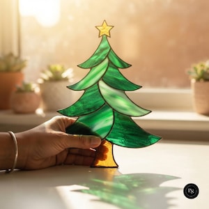 Könnte beinhalten: Ein Weihnachtsbaum-Ornament aus Buntglas mit geschichteten grünen Glaspaneelen, dunklen Lötstellen, einem gelben Glasstern an der Spitze und einem braunen Glasstamm. Das durchscheinende Glas erzeugt einen lebendigen grünen Farbton.
