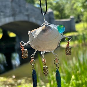 May include: A hanging ornament featuring a central white seashell wrapped in silver wire, accented with a single blue and green glass bead. Three dangling charms include two small fish-shaped pendants and metallic beads suspended by copper wire.