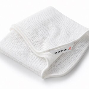 May include: A folded white textile cloth with a ribbed knit texture. A small white fabric label is attached to the edge, featuring the black text NAKAMURAYA and a small red square icon containing a white character symbol.