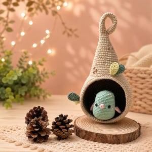 May include: A beige crocheted hanging pod with a pointed top and a dark interior, containing a small, light green crocheted bird figure with black eyes and a pink beak. The pod features small green leaf accents on the exterior surface.