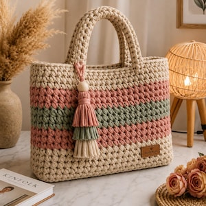 Op de afbeelding: Een rechthoekige gehaakte handtas met horizontale strepen in crème, oudroze en saliegroen. De tas heeft twee stevige handvatten en een decoratieve kwast met een houten kraal. Een klein bruin rechthoekig label met de tekst Hand Made is bevestigd.