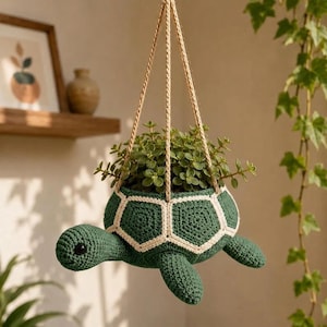 Op de afbeelding: Een gehaakte hangende plantenbak in de vorm van een schildpad in groen en crème, met een gedetailleerd schildpatroon en een centrale opening voor een kleine plant. De schildpad heeft een ronde kop met een zwart oog en vier ledematen, opgehangen aan drie koorden.