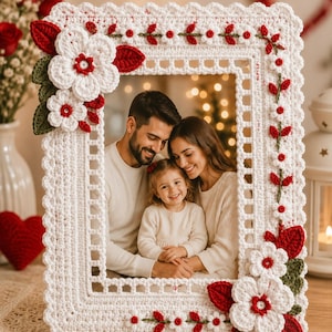 Puede incluir: Un marco de fotos rectangular con un intrincado trabajo de ganchillo blanco y un borde festoneado. El marco está decorado con flores blancas de ganchillo en tres dimensiones con centros rojos, hojas rojas y pequeñas enredaderas verdes.