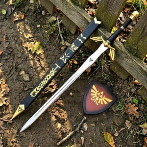 May include: A silver-colored sword with a black handle and gold-toned crossguard, accompanied by a black scabbard featuring gold-colored decorative patterns and a small shield with a gold-colored emblem on a dark red background.