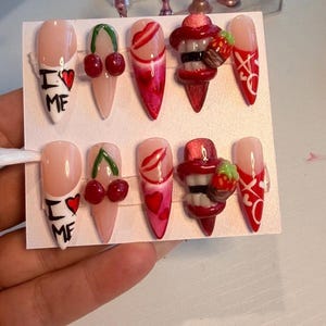 May include: A set of ten stiletto-shaped press-on nails featuring a nude base with red and white designs. Patterns include the text I heart ME, red cherries, red lips, 3D strawberry charms, and white X and O symbols on a red background.
