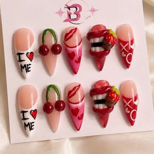 May include: A set of ten stiletto-shaped press-on nails featuring various designs including red cherries, red lips, hearts, white text reading I LOVE ME, and 3D red lips with white teeth and a small strawberry charm, all displayed on a white card.