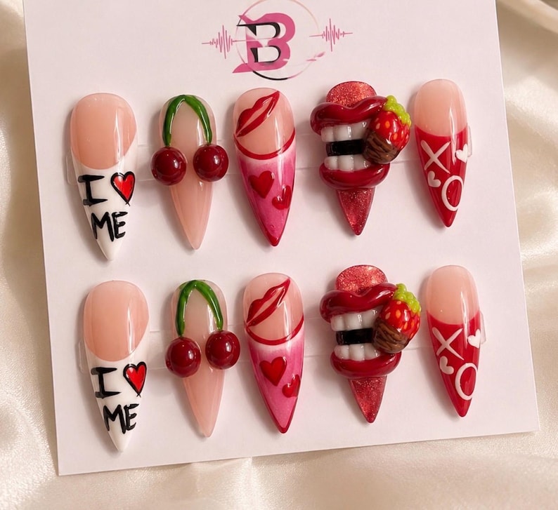 May include: A set of ten stiletto-shaped press-on nails featuring various designs including red cherries, red lips, hearts, white text reading I LOVE ME, and 3D red lips with white teeth and a small strawberry charm, all displayed on a white card.
