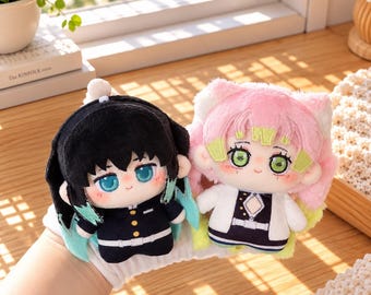 May include: Two plush dolls featuring anime-style characters. The left doll has black hair with teal tips and a dark uniform. The right doll has pink hair with lime green tips, cat ears, and a white and dark uniform. Both have large eyes.