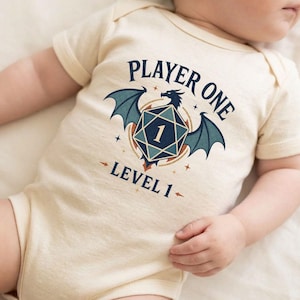 Op de afbeelding: Een crèmekleurige romper voor baby's met een donkerblauwe grafische print. Het ontwerp toont het silhouet van een gevleugelde draak achter een twintigzijdige dobbelsteen met het cijfer 1, omringd door de tekst PLAYER ONE en LEVEL 1 in blauw.