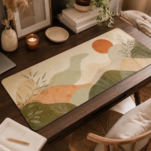 Puede incluir: Una alfombrilla de escritorio rectangular con un diseño de paisaje abstracto que muestra colinas en tonos verde salvia, crema y naranja quemado. La superficie incluye un sol naranja prominente y delicadas ilustraciones lineales de ramas botánicas.
