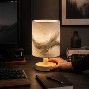 May include: A cylindrical table lamp with a light wood base and a fabric shade featuring a grey and white abstract marbled pattern. The lamp emits a warm glow, illuminating the textured surface of the shade with subtle dark swirls.