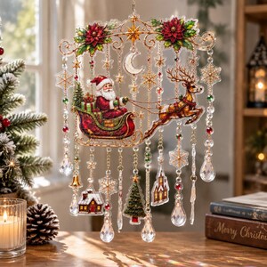 Op de afbeelding: Een decoratieve hangende versiering met een centrale afbeelding van de Kerstman in een arrenslee getrokken door een rendier, versierd met kerststerren, sterren, een maansikkel en bungelende kristallen, sneeuwvlokken, huis en boom.
