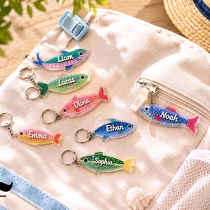 May include: A collection of seven acrylic fish-shaped keychains in various colors including blue, green, pink, and yellow. Each keychain features a white printed name: Liam, Lucas, Olivia, Emma, Ethan, Noah, and Sophia, attached to a metal ring.