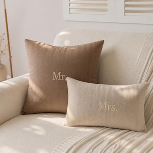 May include: A square brown throw pillow embroidered with the text Mr. and a rectangular cream throw pillow embroidered with the text Mrs. Both pillows feature a textured fabric finish and are positioned together on a light-colored surface.