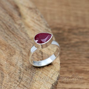 Puede incluir: Un anillo de plata con una piedra preciosa de color rojo intenso en forma de pera, engastada en un bisel de metal liso. La banda es de plata pulida con un sello visible en la superficie interior. El diseño resalta el contraste entre la piedra roja vibrante y el acabado metálico plateado de la estructura del anillo.