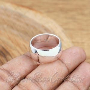 May include: A wide, polished silver band ring with a smooth, reflective surface. The interior of the band features a small, stamped hallmark indicating its metal composition. The ring has a rounded, substantial profile with a high-shine finish.