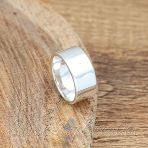 May include: A wide, polished silver band ring with a smooth, reflective surface. The ring features a simple, unadorned design with a high-shine finish that catches the light, showcasing a clean, metallic appearance throughout the entire band.