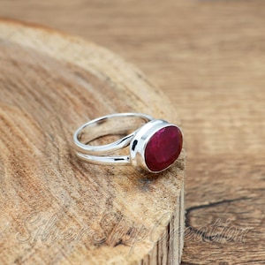 May include: A silver ring featuring a polished, oval-shaped deep red ruby gemstone set in a smooth metal bezel. The band of the ring is split into two distinct silver strands that converge at the base, creating a simple and clean design for the jewellery.