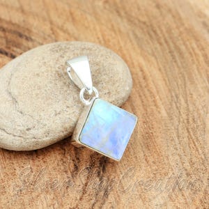 May include: A square-shaped pendant featuring a polished, iridescent moonstone set in a silver frame with a triangular bail at the top. The stone displays a soft blue glow across its surface, reflecting light against the metallic silver setting.