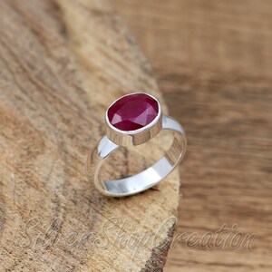 Könnte beinhalten: Ein Silberring mit einem facettierten, ovalen, tiefroten Rubin, der in einer glatten Metallfassung sitzt. Der Ring besteht aus poliertem Silber und der Text Silver Shop Creation ist im unteren Teil des Bildes zu sehen.