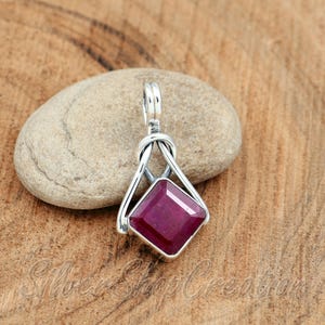 May include: A silver pendant featuring a square-cut, deep magenta-red gemstone set in a polished metal frame. The design includes a looped bail at the top, connecting to a triangular structure that holds the faceted stone securely in place.