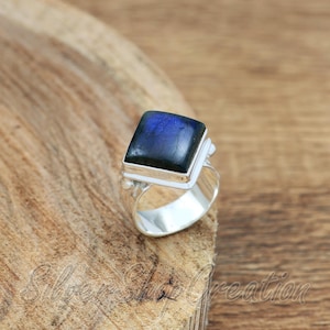 May include: A silver ring featuring a square-cut labradorite gemstone with a dark blue iridescent sheen. The stone is set in a smooth, polished silver bezel atop a wide, solid silver band with a textured, hammered finish on the sides.