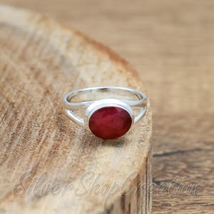 Può includere: Un anello in argento con una pietra preziosa rosso scuro di forma ovale, lucidata e incastonata in una montatura in metallo liscio. Il gambo è realizzato in un sottile filo d'argento lucido che si divide in due verso l'incastonatura.