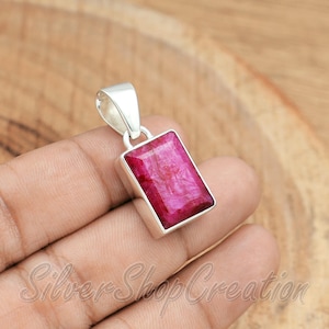 Puede incluir: Un colgante rectangular con una piedra preciosa facetada de color rosa intenso, engastada en un marco de plata pulida con una anilla de plata en la parte superior. La piedra tiene un aspecto natural texturizado con inclusiones visibles.