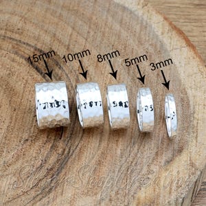 May include: Five silver-colored rings with a hammered texture, arranged in descending order of width. The rings are labeled with arrows indicating widths of 15mm, 10mm, 8mm, 5mm, and 3mm, showcasing the variation in size for these metallic bands.