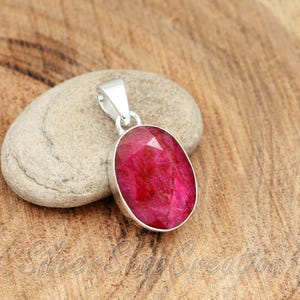 Puede incluir: Un colgante de forma ovalada con una piedra preciosa facetada de color rojo rosado intenso, engastada en un bisel de plata pulida. El colgante incluye una reasa de plata en la parte superior. El texto Silver Shop Creation es visible abajo.