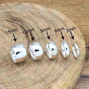 Könnte beinhalten: Fünf polierte Silberringe in einer Reihe angeordnet, mit Breiten von 3mm bis 15mm. Jeder Ring ist mit seiner spezifischen Breitenangabe beschriftet, darunter 3mm, 6mm, 9mm, 12mm und 15mm, markiert durch schwarze Pfeile auf jeden Ring.