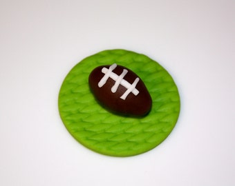 May include: A brown football with white stripes rests on a green, textured, circular base. The base resembles a patch of grass. The image is set against a plain white background, highlighting the football and its base.