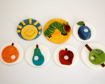 May include: Six decorated cupcakes with a white background. The cupcakes are decorated with a smiling sun, a green caterpillar, a blue blueberry, a red apple, a green pear, and a green leaf.