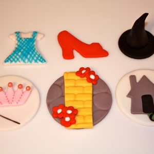 May include: Six fondant cupcake toppers featuring characters and objects from the Wizard of Oz. The toppers include Dorothy's blue gingham dress, ruby slippers, the Wicked Witch's hat, a yellow brick road, a crown, and a grey house.