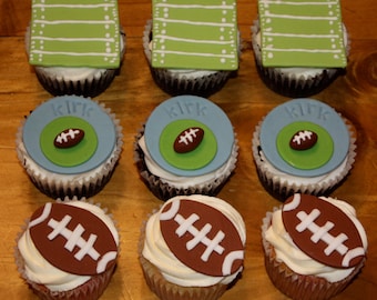May include: Nine cupcakes with white frosting. Three cupcakes have green and white football field toppers. Three cupcakes have blue and green toppers with a football and the name "kirk". The remaining three cupcakes have football-shaped toppers.