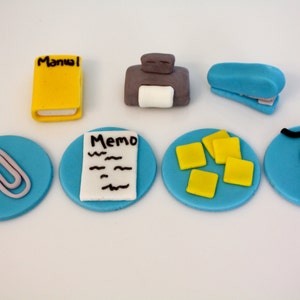 May include: Six blue and white cupcake toppers with office-themed decorations. The toppers feature a yellow book with the word "Manual" on the cover, a gray printer, a blue stapler, a paperclip, a white memo pad with the word "Memo" on it, yellow sticky notes, and a black computer mouse.
