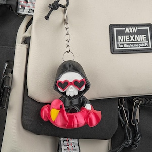 Peut inclure: Un porte-clés représentant une figurine de crâne blanc portant une cape à capuche noire et des lunettes de soleil en forme de cœur rouge, assise dans une bouée flamant rose. Le porte-clés est attaché à un sac à dos beige et noir avec l'étiquette NXN NIEXNIE.