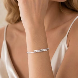 May include: A silver-toned bypass bracelet featuring a continuous row of small, clear, sparkling stones set along the entire length of the band. The design consists of two overlapping ends, each finished with a small, smooth, round metal bead.